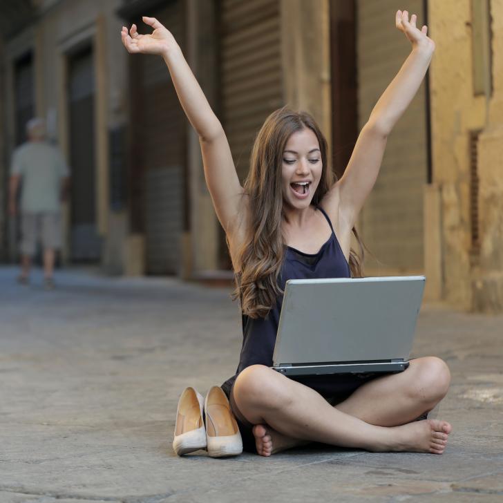 Eine junge Frau sitzt mit ihrem Laptop am Schoß auf einer Straße und wirft ihre Arme vor Freude in die Höhe